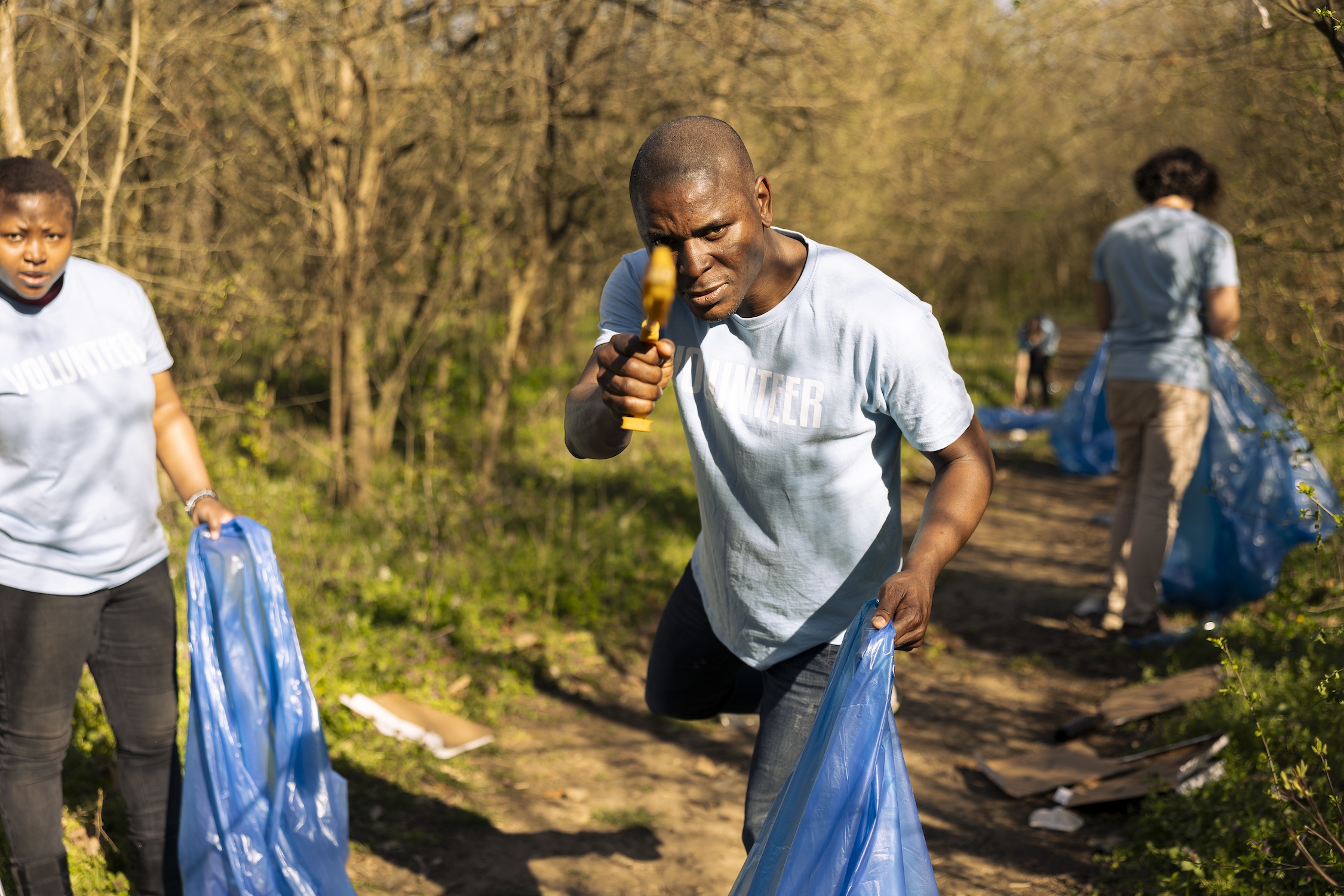 Male volunteer having fun cleaning the forest from garbage, using a long claw tool to grab litter and plastic waste. Tidying the woodland to conserve natural ecosystem, save the planet.